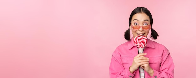 Tonto y lindo modelo femenino asiático lamiendo piruleta comiendo caramelos dulces y sonriente mirando emocionado de pie sobre fondo de color rosa