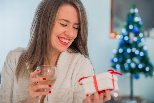 Toothy sonriente mujer con labios rojos celebración de caja de regalo y vid de vid. Mujer feliz celebración de caja de regalo. Bebida de champán. Vid de vid.