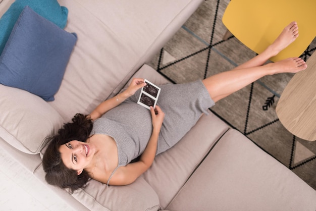 Top-Ansicht einer jungen schwangeren Frau, die nach dem Ultraschallfoto des Babys sucht, während sie sich zu Hause auf dem Sofa entspannt