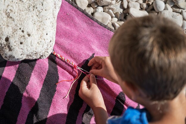 Foto top-ansicht eines jungen, der ein stringarmband an einem kiesstrand knüpft