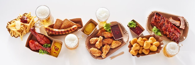 Top-Ansicht von Glasbechern mit Lagerbier, die auf einem weißen Tisch mit verschiedenen Fleisch und Fastfood im Studio serviert werden
