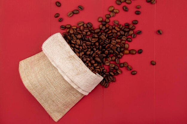 Top-Aufnahme von frisch gerösteten Kaffeebohnen, die von einem Burlap-Beutel auf einem roten Hintergrund herausfallen