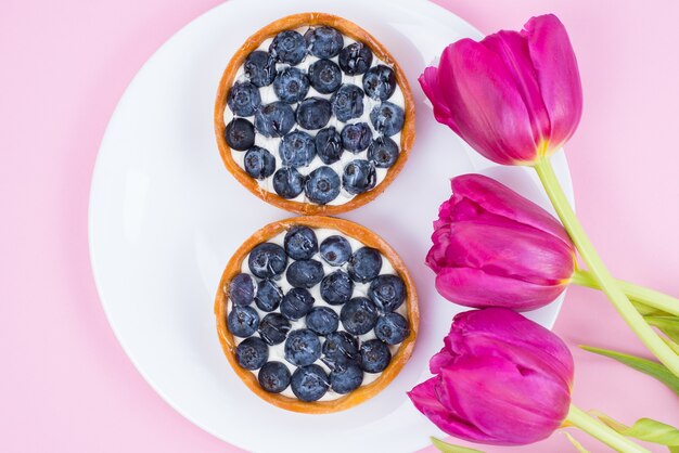 Top Flat Lay Flatlay über Ansicht Foto von schönen hellen Blumen und zwei köstlichen Törtchen mit süßen Blaubeeren isoliert Pastell weiche Farbe Oberfläche