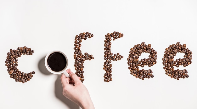 Top-View von Wortkaffee aus Kaffeebohnen und menschlicher Hand, die eine Tasse schwarzer Kaffee hält