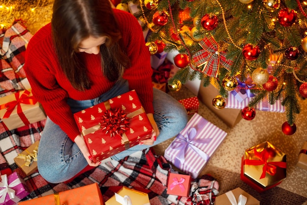 Topdown-Aufnahme eines Mädchens, das unter einem geschmückten Weihnachtsbaum sitzt, mit einem Geschenk-Weihnachtsfeierkonzept