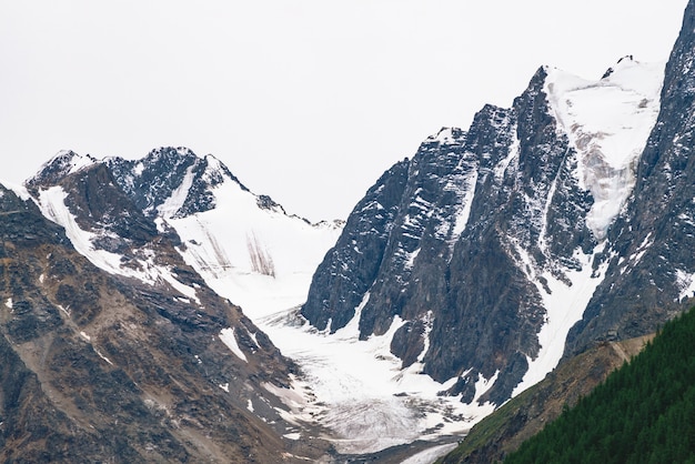 Topo da montanha de neve atrás da colina com floresta sob céu nublado. Cume rochoso em tempo nublado. Branca de neve na geleira. Paisagem atmosférica de natureza majestosa.