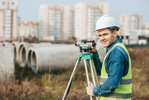 Foto topógrafo com nível digital sorrindo para a câmera com materiais de construção ao fundo