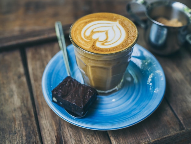 Topping de café con leche Piccolo con arte floral de la leche en un vaso pequeño con un trozo de pastel casero de brownies en un plato de cerámica azul sobre una mesa de madera