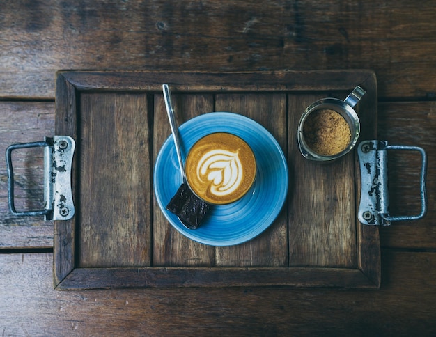 Topping de café con leche Piccolo con arte floral de la leche en un vaso pequeño con un trozo de pastel casero de brownies en un plato de cerámica azul sobre una mesa de madera