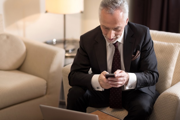 Un toque. Sonriente hombre de negocios de edad alegre sosteniendo el teléfono y sentado en el hotel frente a la computadora portátil mientras trabaja en el proyecto y expresa entusiasmo