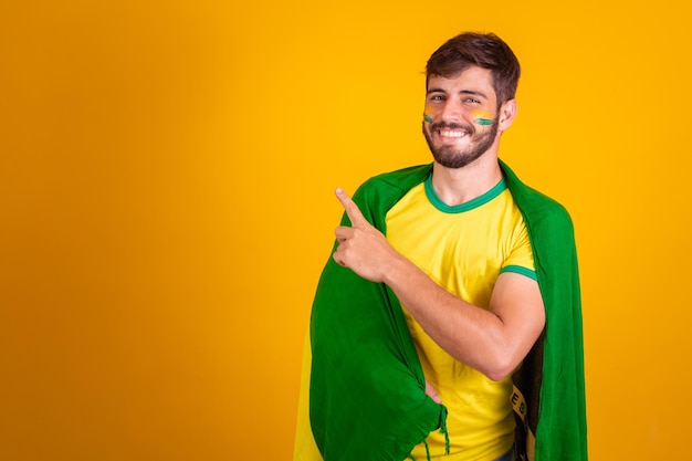 Foto torcedor brasileiro com uma bandeira do brasil nas costas em fundo amarelo, apontando para o espaço para texto