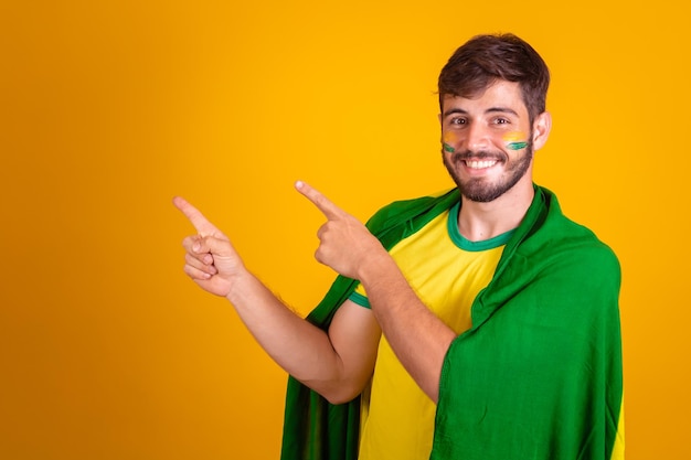 Torcedor brasileiro com uma bandeira do brasil nas costas em fundo amarelo, apontando para o espaço para texto