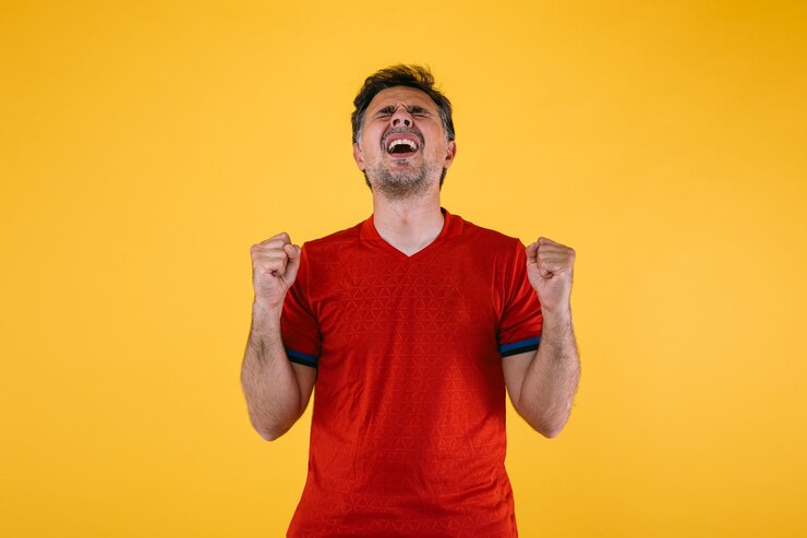 Torcedor de futebol com uma camisa vermelha cerra os punhos e grita torcendo por seu time.