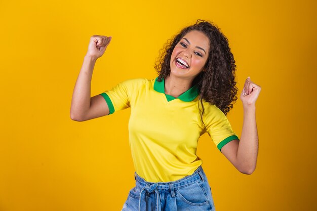 Foto torcedor do brasil. fã de mulher brasileira de cabelo encaracolado comemorando no futebol, jogo de futebol em fundo amarelo. cores do brasil.