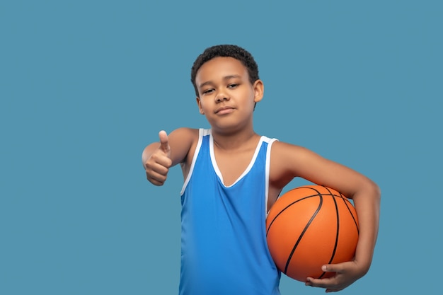 Torne-se profissional. Menino alegre de escola primária de pele escura com uma bola mostrando um gesto de ok parado sobre um fundo azul