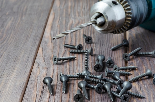 Tornillos de metal y un taladro eléctrico sobre una mesa de madera. Herramienta de fijación y reparación.