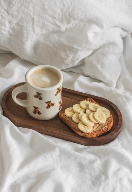 Torrada com pasta de amendoim e cappuccino de banana em uma bandeja de madeira em uma cama com roupa de cama de linho branco