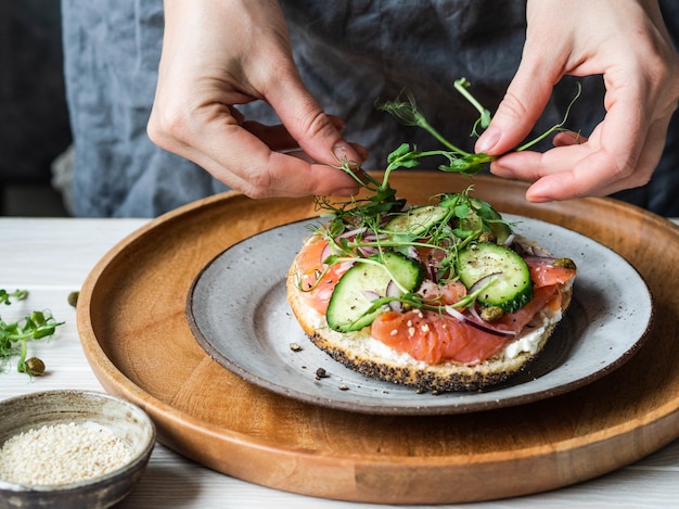 Torrada saudável com creme de queijo, salmão, pepino fresco, alcaparras, cebola roxa, pimenta preta ee uma mulher incerta coloca em brotos de ervilha sanduíche.