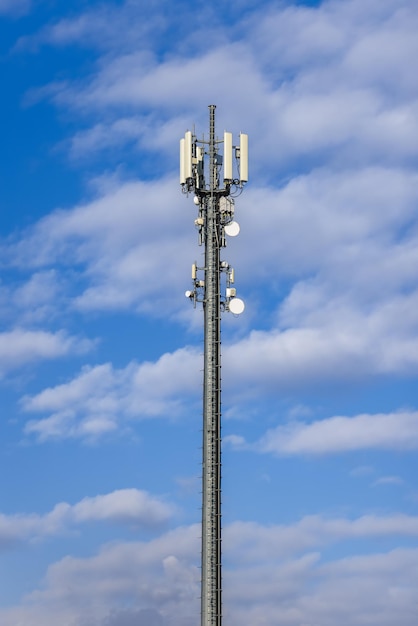 Torre de celulares con varias antenas y amplificadores de señal contra un cielo azul con nubes Foto vertical