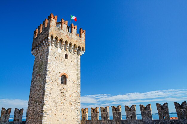 Foto torre com bandeira italiana e parede de defesa de pedra com merlons do castelo de scaligero castello di sirmione