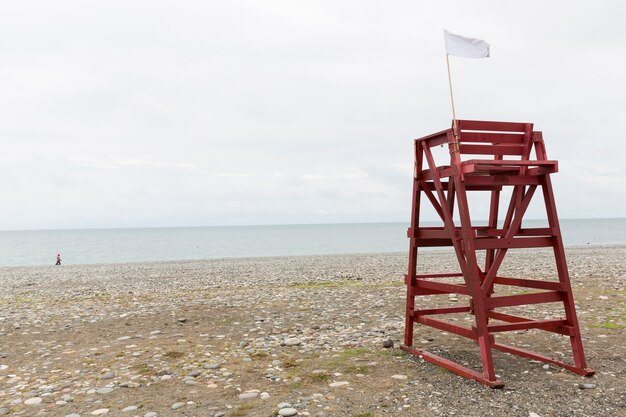 La torre roja de salvavidas en la playa de guijarros de la ciudad turística está fuera de temporada. No hay gente, playa vacía. Invierno, nubes. Montañas al fondo. Foto de alta calidad