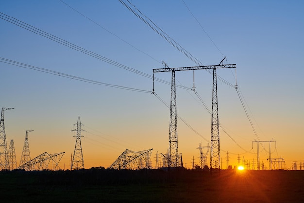 Torres de eletricidade de alta tensão em campo ao pôr do sol e céu azul claro Silhuetas escuras de repetir linhas de energia no nascer do sol laranja Rede de transmissão e distribuição de geração de eletricidade