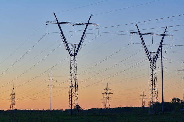 Torres de electricidad de alto voltaje en el campo al atardecer y cielo azul claro Siluetas oscuras de líneas eléctricas repetidas en el amanecer naranja Red de transmisión y distribución de generación de electricidad