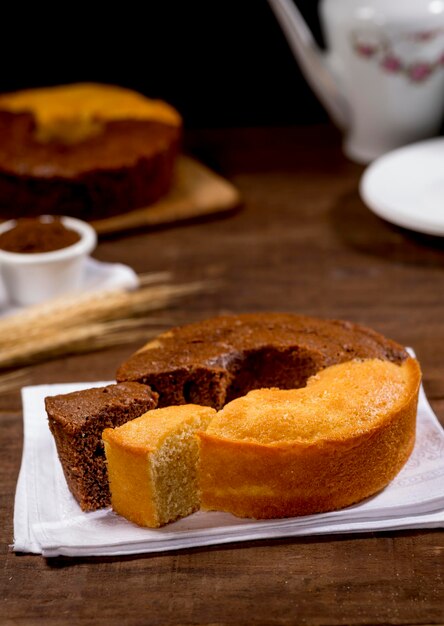 Foto torta de chocolate y harina de trigo y rebanada sobre tela sobre madera con torta en el fondo con rama de trigo, plato blanco, botín blanco al lado.