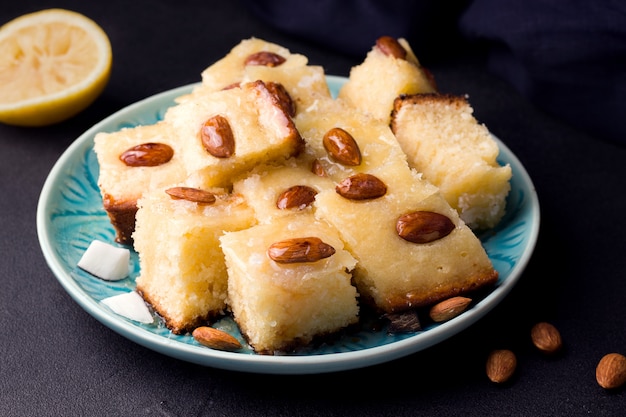 Torta doce árabe basbus cortada em fatias sobre um fundo preto ao lado de um limão e tecido azul