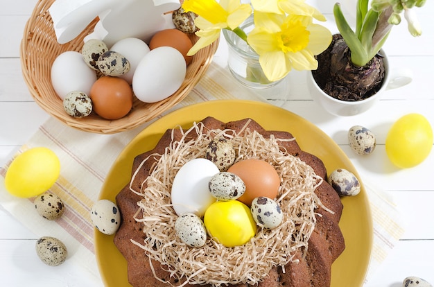 Torta de Pascua con huevos teñidos en un nido, narcisos y jacintos en una taza sobre un fondo de madera rústico blanco