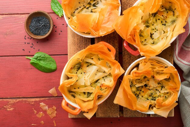 Tortas Filo com queijo feta macio e espinafre em moldes de cerâmica em antigos fundos de mesa de madeira vermelha Tortas Filo porções Tortas Spanakopita pequenas assadas Vista superior Espaço para cópia Cozinha grega tradicional