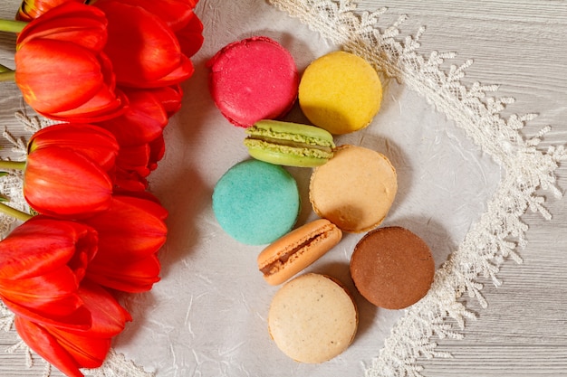 Tortas de macarons de diferente color en servilleta de seda con ramo de tulipanes rojos frescos con fondo de madera. Vista superior