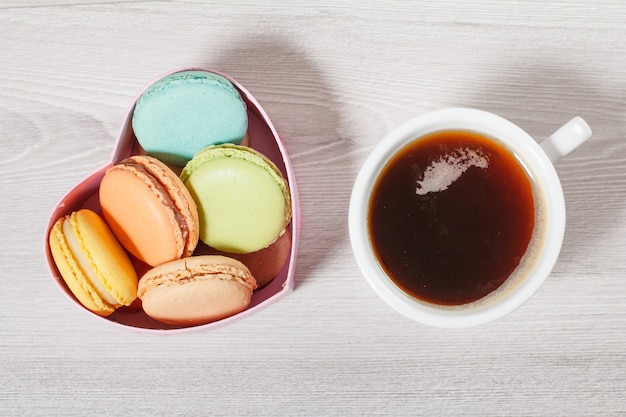 Tortas de macarrones sabrosos de diferente color en caja de cartón en forma de corazón y taza de café en tableros grises. Vista superior