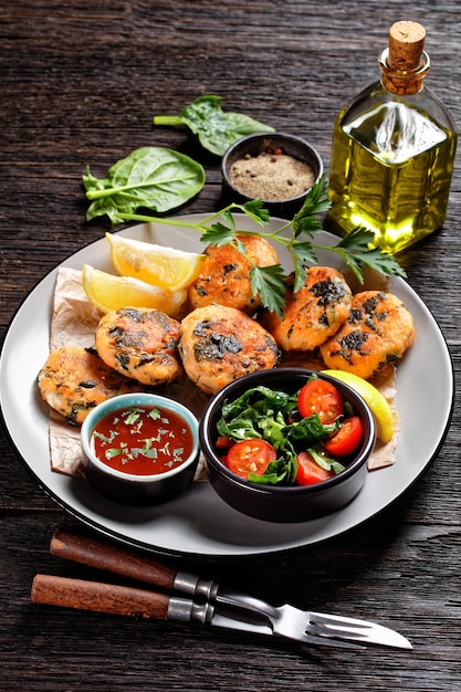 Tortas de pescado de salmón y espinacas con ensalada de verduras frescas, rodajas de limón y salsa de chile dulce en un plato sobre una mesa de madera, vista vertical