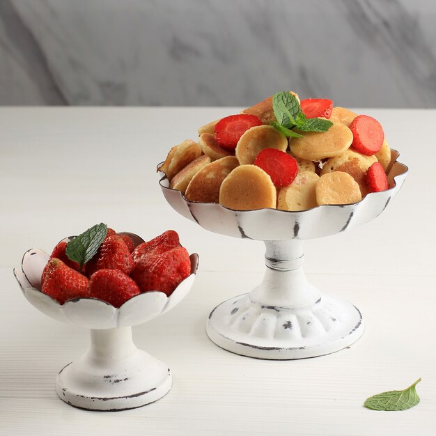 Tortenständer mit winzigen Pfannkuchen-Müsli und Erdbeeren, garniert mit Minzblättern und Zitronenscheibe auf weißem Hintergrund. Trendiges Essen. Mini-Getreidepfannkuchen. Quadratisches Bild mit Marmorhintergrund