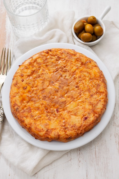 Tortilla de comida tradicional española con patatas en un plato blanco y aceitunas en un tazón pequeño sobre fondo de cerámica.