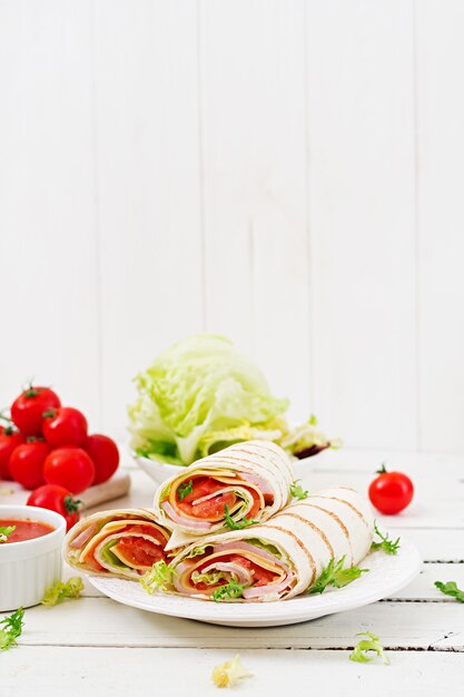 Tortilla envuelta con jamón, queso y tomates en una mesa de madera blanca