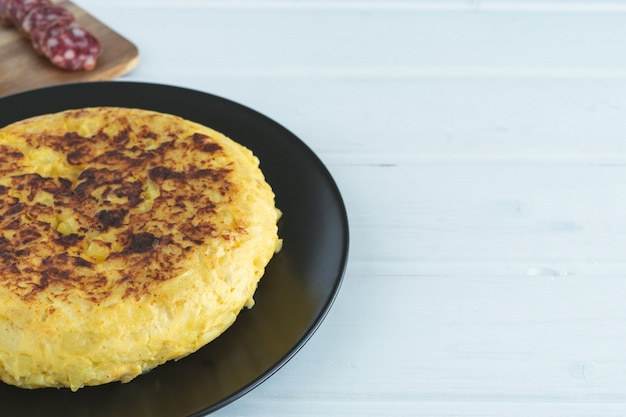 Foto tortilla española en una placa negra sobre un fondo blanco de madera.