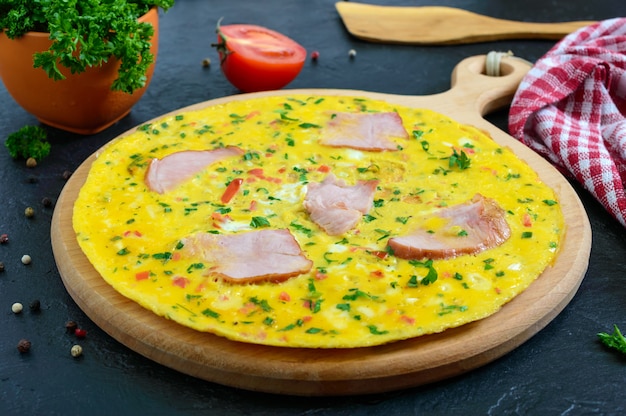 Tortilla con jamón, queso, verduras sobre una tabla de madera sobre un fondo negro. Fritata: un desayuno delicioso y saludable.