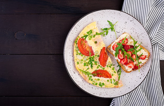 Tortilla con tomate, jamón, cebolla verde y sándwich con fresa en la mesa oscura, vista superior