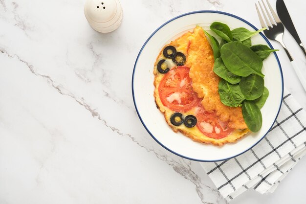 Tortilla con tomates, aceitunas negras, requesón y hierbas de espinacas verdes en un plato blanco sobre fondo de hormigón gris Delicioso desayuno Comida saludable para el desayuno Vista superior