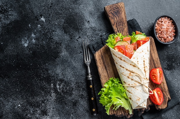 Tortilla Wrap roll de salmón con ensalada, verduras. Fondo negro. Vista superior. Copie el espacio.