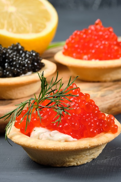 Tortinhas com caviar vermelho e preto em uma placa de madeira com limão e ervas.