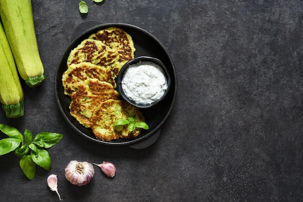 Tortitas de calabacín con salsa y ajo en una sartén sobre un fondo negro. Vista desde arriba.