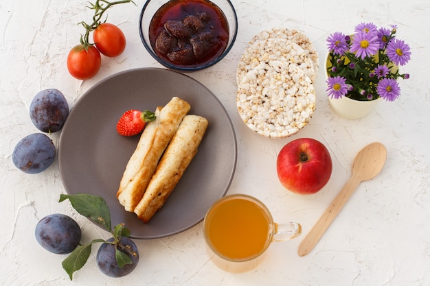 Tortitas caseras rellenas de requesón, una fresa en el plato, ciruelas, un cuenco de cristal con mermelada, una cuchara de madera, manzanas, tortas de arroz inflado, una taza de té y flores. Vista superior.