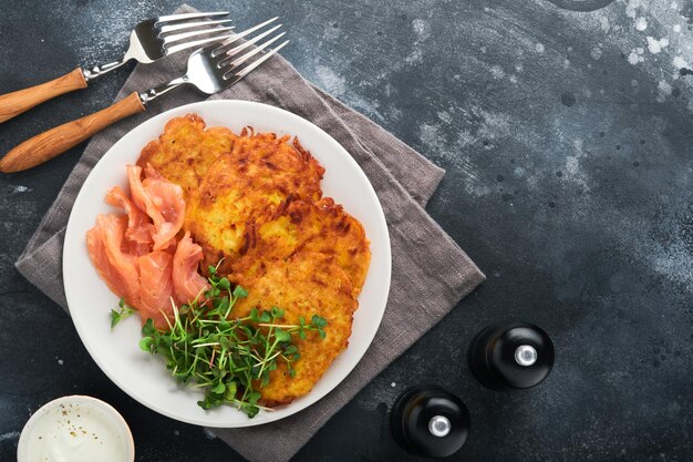 Tortitas de patata Tortitas de patata caseras fritas o latkes con cebollas verdes crema microgreens salmón rojo y salsa en plato rústico sobre fondo de mesa negro antiguo Estilo rústico Vista superior