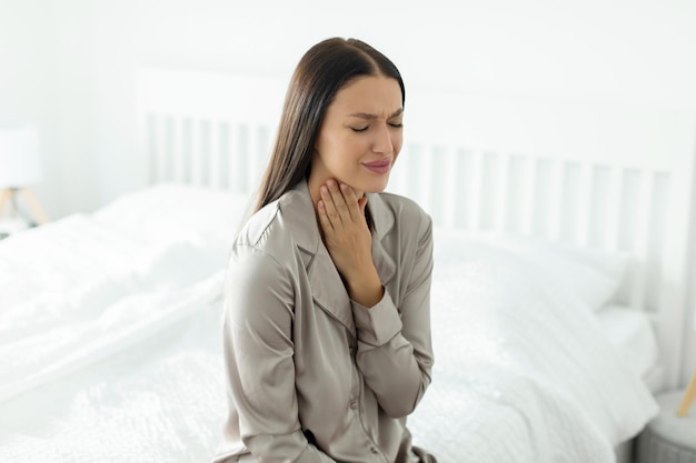 Tose cosquillas mujer joven enferma tocando el cuello dolor de garganta en casa sentado en el borde de la cama resfriado estacional y gripe