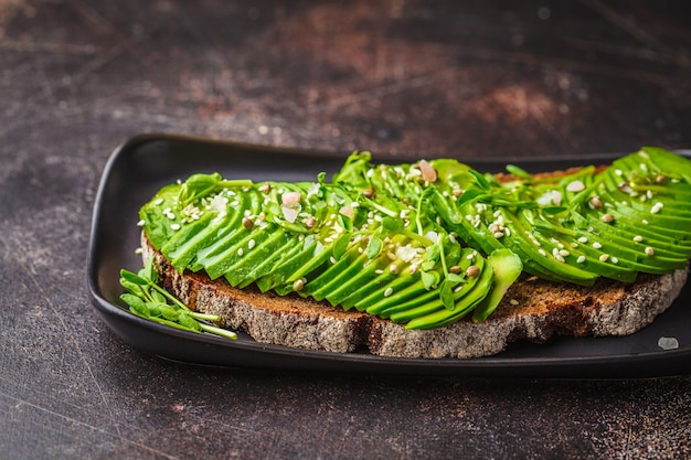 Tostada del aguacate con pan y semillas de centeno en la placa negra, fondo oscuro.