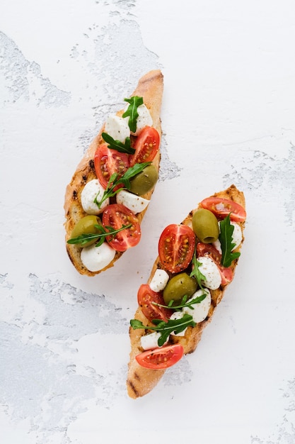 Tostadas de bruschetta Caprese con tomates cherry, mozzarella, aceitunas y albahaca en la superficie de luz vieja placa blanca. Vista superior.