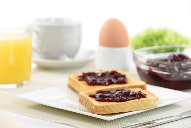 Tostadas crujientes con mermelada de frambuesa en un plato y un huevo pasado por agua junto al jugo de naranja y una taza de café y manzanas sobre fondo blanco.